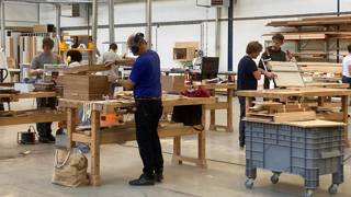 Mensen werken aan houten meubels in een ruime werkplaats met grote werkbanken, gereedschap en stapels hout op rekken.