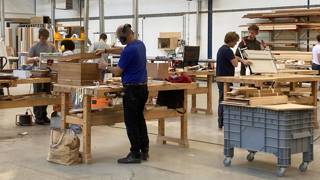 Mensen werken aan houten meubels in een ruime werkplaats met grote werkbanken, gereedschap en stapels hout op rekken.