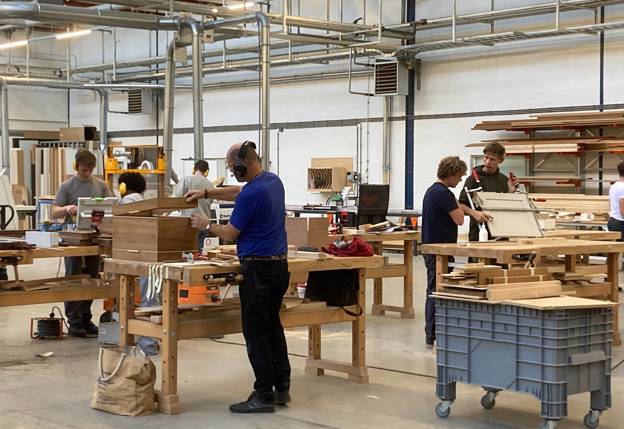 Mensen werken aan houten meubels in een ruime werkplaats met grote werkbanken, gereedschap en stapels hout op rekken.