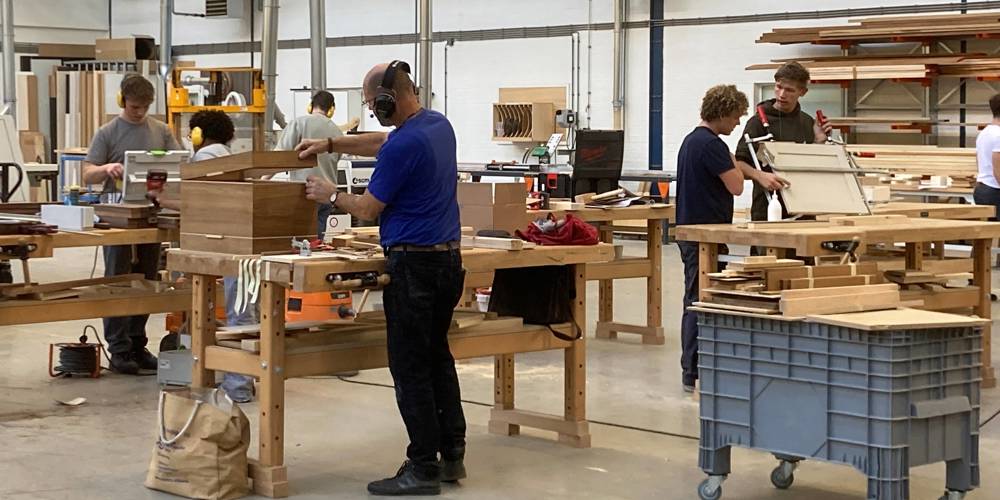 Mensen werken aan houten meubels in een ruime werkplaats met grote werkbanken, gereedschap en stapels hout op rekken.