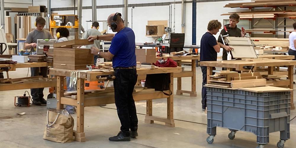 Mensen werken aan houten meubels in een ruime werkplaats met grote werkbanken, gereedschap en stapels hout op rekken.