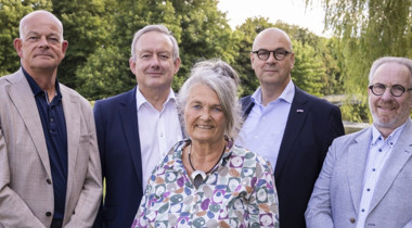 Vijf personen buiten, met bomen en water op de achtergrond.
