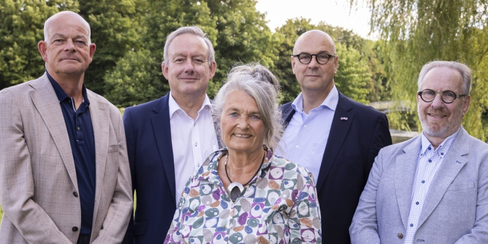 Vijf personen buiten, met bomen en water op de achtergrond.