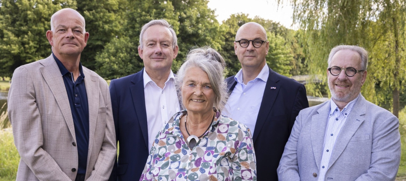 Vijf personen buiten, met bomen en water op de achtergrond.