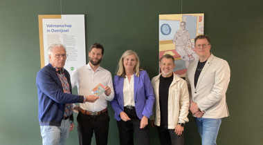 Vijf personen poseren voor een groene wand met een informatiepaneel en een kunstwerk.