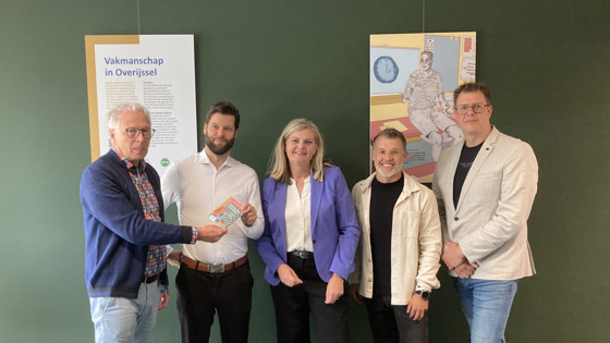 Vijf personen poseren voor een groene wand met een informatiepaneel en een kunstwerk.