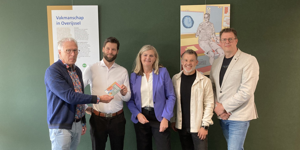 Vijf personen poseren voor een groene wand met een informatiepaneel en een kunstwerk.