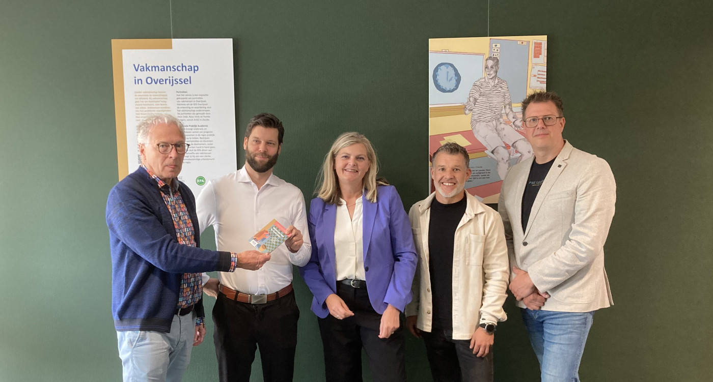 Vijf personen poseren voor een groene wand met een informatiepaneel en een kunstwerk.