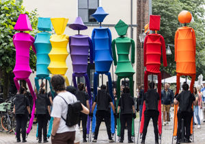  Straatvoorstelling met kleurrijke figuren op stelten, ondersteund door begeleiders in zwart, in een stedelijke omgeving.