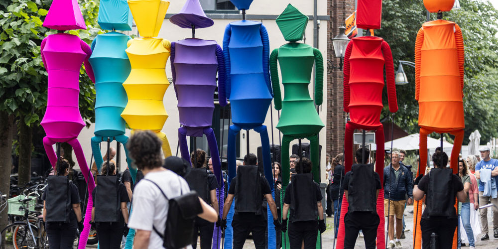  Straatvoorstelling met kleurrijke figuren op stelten, ondersteund door begeleiders in zwart, in een stedelijke omgeving.