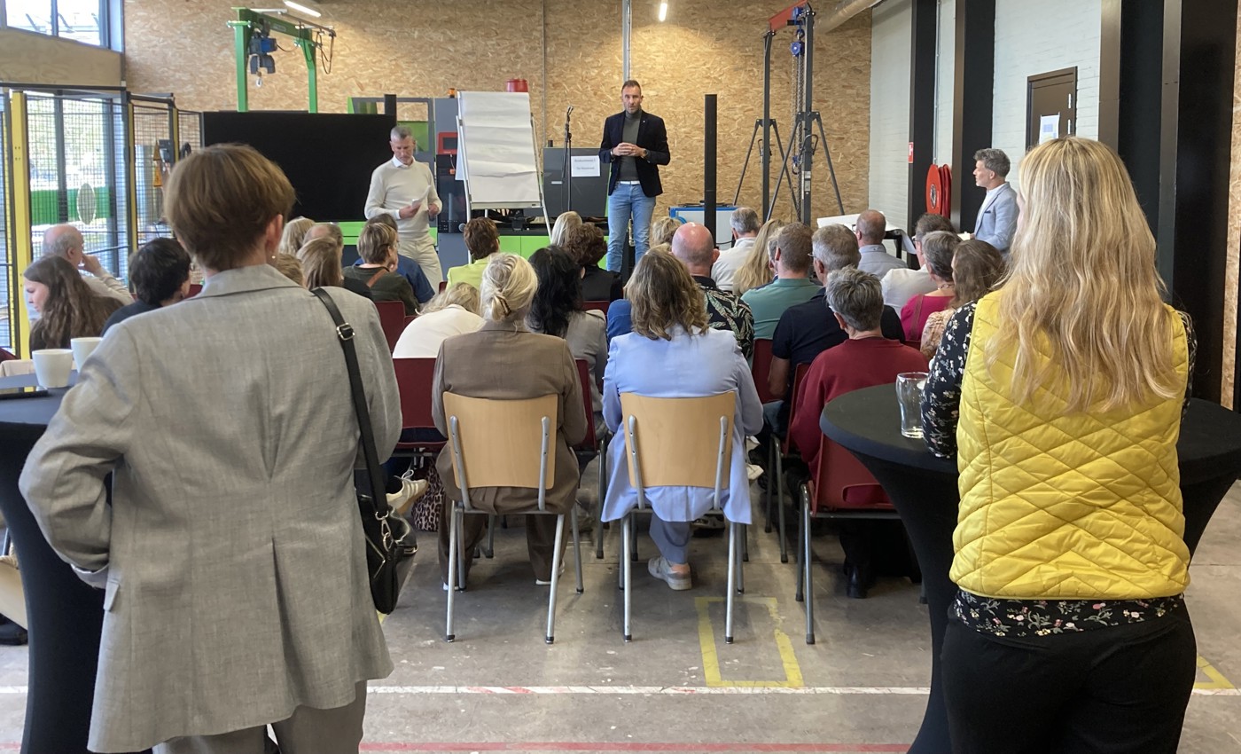 De bijeenkomst in een zaal van de Regionale Praktijk Academie met het publiek vanaf de rug gezien. De meesten luisteren zittend naar een spreker op het podium. Vooraan in beeld staan 2 aanwezigen te luisteren die we ook op de rug zien.