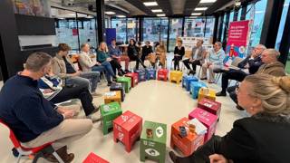 Mensen zitten in een kring in gesprek, met gekleurde SDG-blokken in het midden.