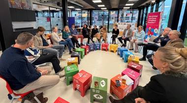 Mensen zitten in een kring in gesprek, met gekleurde SDG-blokken in het midden.