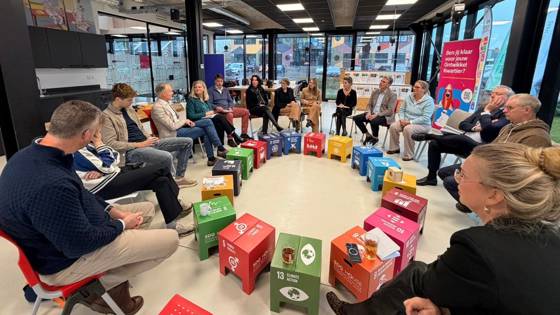 Mensen zitten in een kring in gesprek, met gekleurde SDG-blokken in het midden.