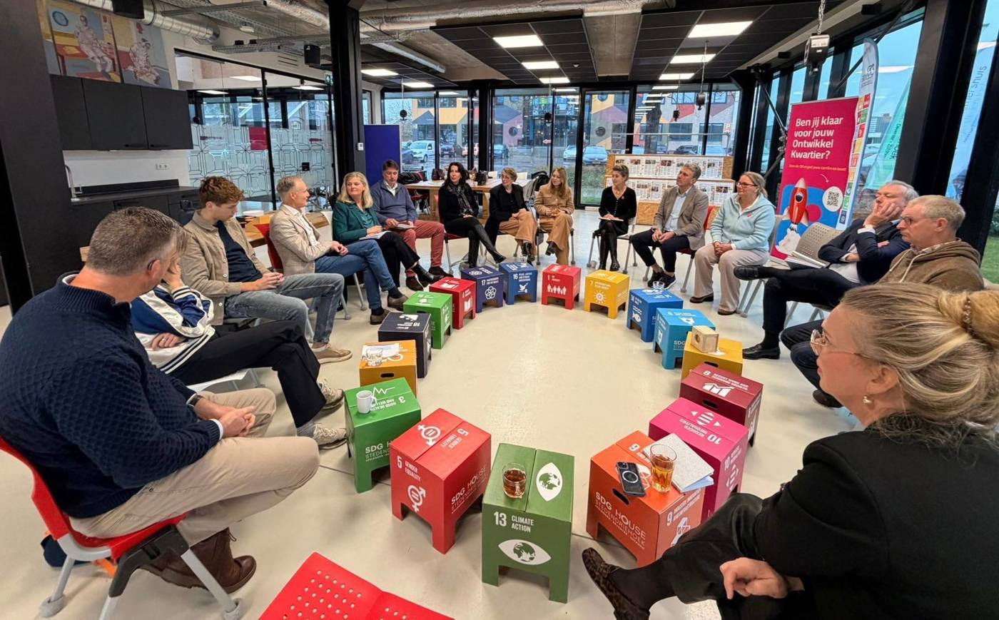 Mensen zitten in een kring in gesprek, met gekleurde SDG-blokken in het midden.