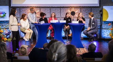 Paneldiscussie tijdens een symposium, met meerdere personen rond drie blauwe statafels op een podium, publiek op de voorgrond.