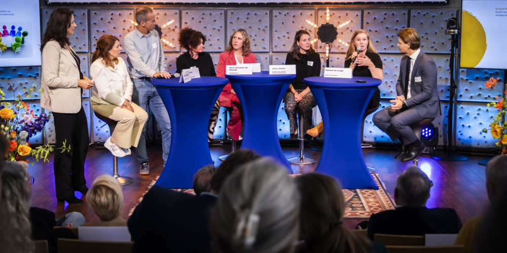 Paneldiscussie tijdens een symposium, met meerdere personen rond drie blauwe statafels op een podium, publiek op de voorgrond.