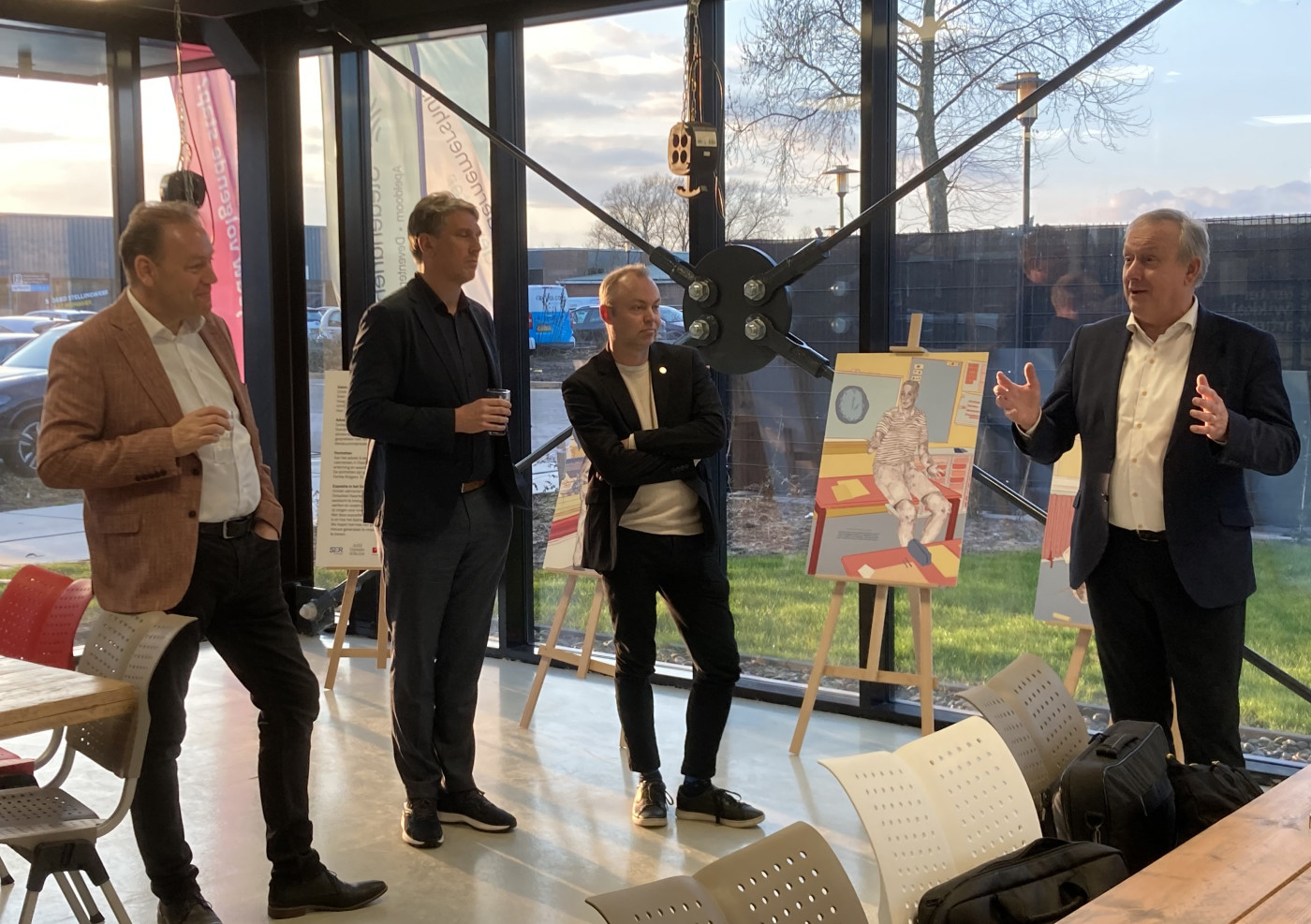 Vier personen in gesprek in een moderne ruimte met grote ramen en kunstwerken op ezels.