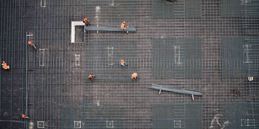 Fotograaf: Saad Salim (Unsplash) - Luchtfoto van bouwplaats met arbeiders in oranje hesjes op een stalen wapeningsnet voor een betonnen fundering.