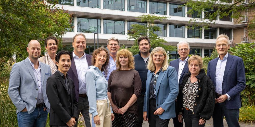 Groepsfoto van meerdere personen staand in een tuinachtige omgeving met groen en bomen, voor een modern kantoorgebouw met grote ramen.