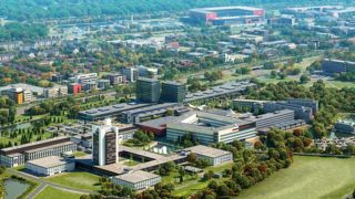 Luchtfoto van diverse gebouwen, parkeerplaatsen, groen en een stadion op de achtergrond.