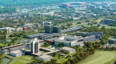 Luchtfoto van diverse gebouwen, parkeerplaatsen, groen en een stadion op de achtergrond.