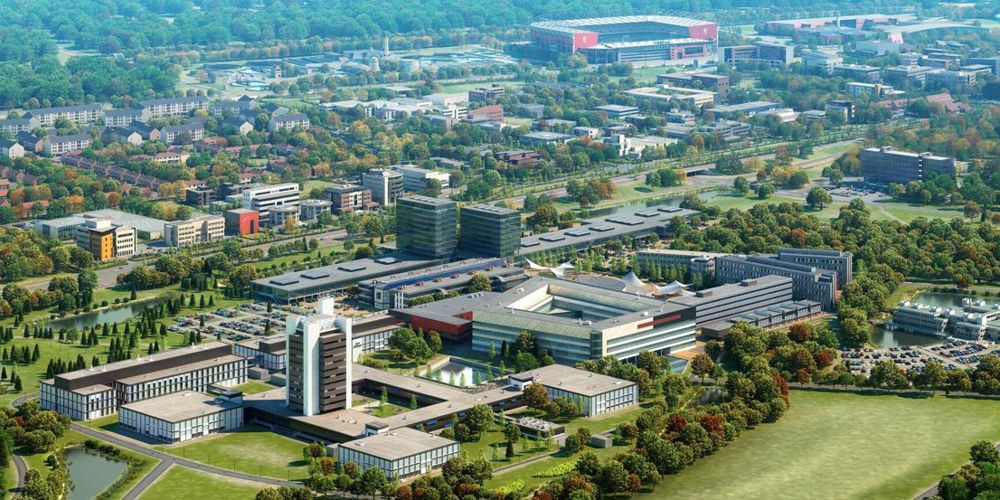Luchtfoto van diverse gebouwen, parkeerplaatsen, groen en een stadion op de achtergrond.