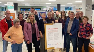 Een groep mensen staat in een bibliotheek rondom een bord op een ezel met de titel “Vakmanschap” en tekst daaronder. Op de achtergrond zijn boekenrekken en een klok aan de muur zichtbaar.