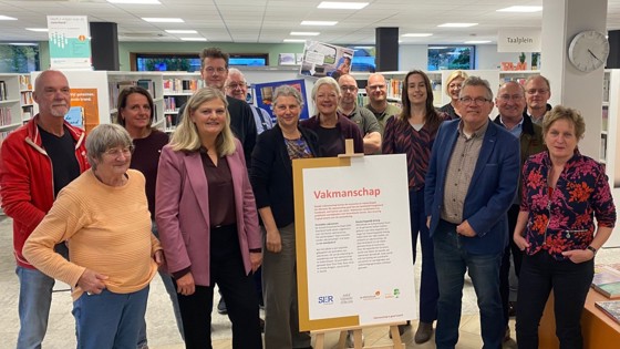 Een groep mensen staat in een bibliotheek rondom een bord op een ezel met de titel “Vakmanschap” en tekst daaronder. Op de achtergrond zijn boekenrekken en een klok aan de muur zichtbaar.