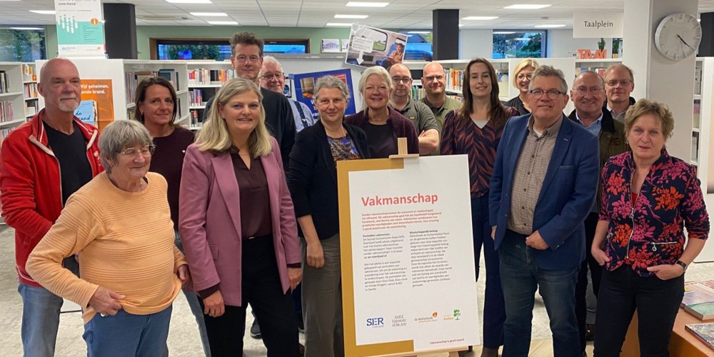 Een groep mensen staat in een bibliotheek rondom een bord op een ezel met de titel “Vakmanschap” en tekst daaronder. Op de achtergrond zijn boekenrekken en een klok aan de muur zichtbaar.